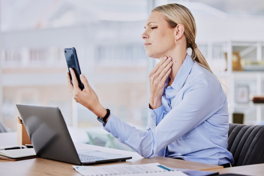 Woman looking at neck lines and wrinkles on phone screen while using computer